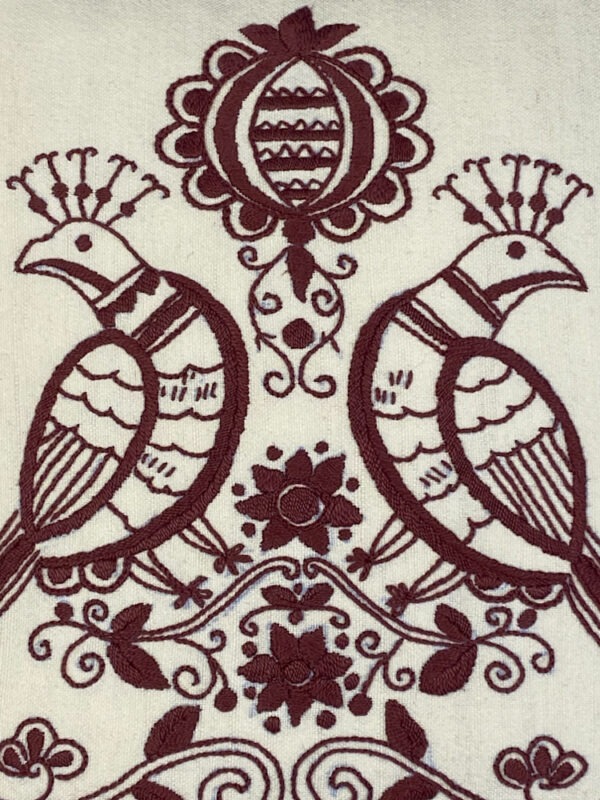 Detailansicht Stickerei, Sofakissen aus weißem Leinen, bestickt mit zwei Vögeln, Rücken an Rücken, und einer Blumengirlande, kleinen Blüten und einem Emblem in dunkelrot.